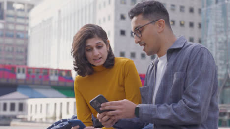 Mid-Shot-of-Two-Students-Talking-and-Using-Smartphone-with-City-Backdrop