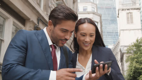 Empresario-Y-Empresaria-Fuera-De-Las-Oficinas-De-La-Ciudad-De-Londres-Celebrando-Buenas-Noticias-En-El-Teléfono-Móvil