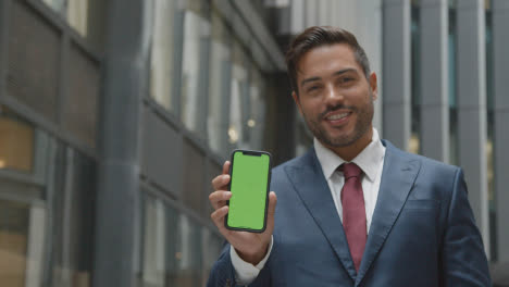Portrait-Of-Businessman-Outside-City-Of-London-Offices-Holding-Up-Green-Screen-Mobile-Phone