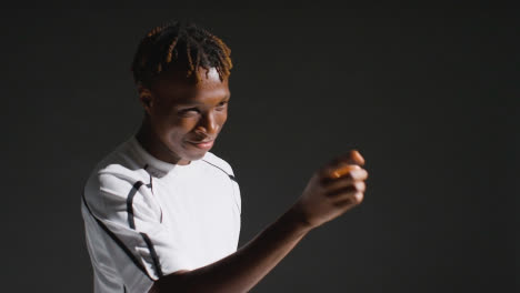 Low-Key-Studio-Portrait-Of-Young-Male-Footballer-Wearing-Club-Kit-Celebrating-Goal-Pretending-To-Shoot-Bow-And-Arrow