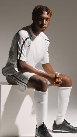 Vertical-Video-Studio-Portrait-Of-Young-Male-Footballer-Wearing-Club-Kit-Sitting-On-Block