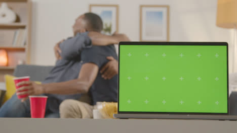 Two-Men-Celebrating-Watching-Sports-Game-On-TV-At-Home-With-Green-Screen-Laptop-In-Foreground-