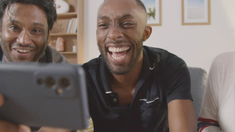 Multi-Cultural-Friends-Celebrating-Watching-Sports-Game-On-Mobile-Phone-Sitting-On-Sofa-At-Home-Together-2