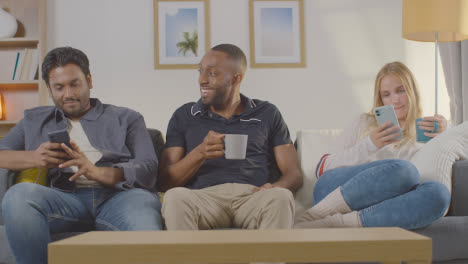 Couple-Sitting-On-Sofa-At-Home-Looking-At-Their-Mobile-Phones-Instead-Of-Talking-To-Friend