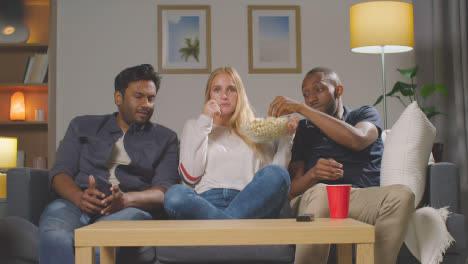 Multi-Cultural-Friends-Sitting-On-Sofa-At-Home-Watching-TV-Movie-And-Throwing-Popcorn-