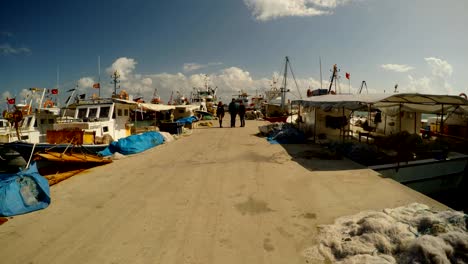 three-people-on-a-sea-fishing-Mediterranean-pier-with-boats,-a-time-lapse-in-a-strong-wind-on-a-sunny-day