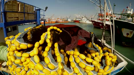 trawl-fishing-net-with-yellow-floats-lies-on-the-boat,-ships-at-the-pier-in-the-distance