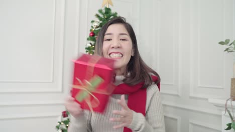 Cheerful-happy-young-Asian-woman-holding-christmas-gifts-smiling-to-camera-in-her-living-room-at-home-in-Christmas-Festival.-Lifestyle-woman-celebrate-Christmas-and-New-year-concept.