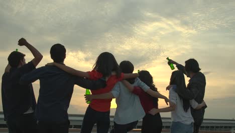 Group-of-young-friends-dancing-and-having-fun-celebrate-with-toast-and-clinking-raising-glasses-at-summer-rooftop-party-with-sunset-back-ground.-Slow-Motion.