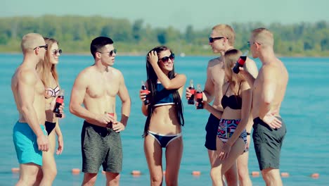 Young-happy-friends-stand-in-a-semicircle-and-communicate-on-the-beach-near-the-water-with-soda-bottles