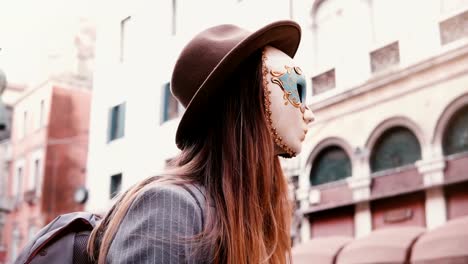 Woman-with-long-hair-and-hat-wearing-a-white-carnival-face-mask-in-Venice-street,-Italy-looking-at-camera,-turning-back.