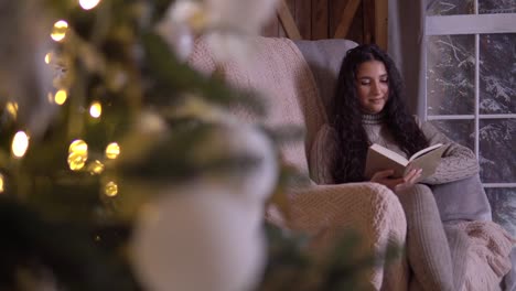 Una-joven-se-sienta-en-una-silla-cerca-del-árbol-de-Navidad-y-lee-un-libro