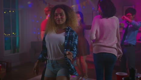 En-la-salvaje-fiesta-de-casa:-hermosa-chica-negra-baila-seductoramente-en-luces-de-neón.-En-el-fondo-otras-personas-divirtiéndose,-Clubbing.