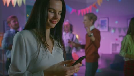 Auf-die-wilde-Hausparty:-schönes-junges-Mädchen-nutzt-Smartphone,-blättert-durch-Internet-und-Social-Network.-In-der-Hintergrund-Menge-junger-Menschen-aus-tanzen-und-Spaß-haben.