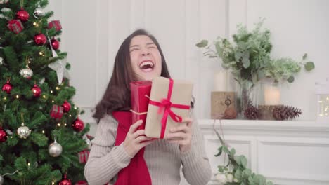 Cheerful-happy-young-Asian-woman-holding-christmas-gifts-smiling-to-camera-in-her-living-room-at-home-in-Christmas-Festival.-Lifestyle-woman-celebrate-Christmas-and-New-year-concept.