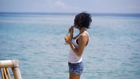 A-young-girl-dressed-in-a-white-T-shirt-and-short-denim-shorts-standing-next-to-the-blue-ocean-and-the-fun-is-drinking-orange-juice-through-a-straw