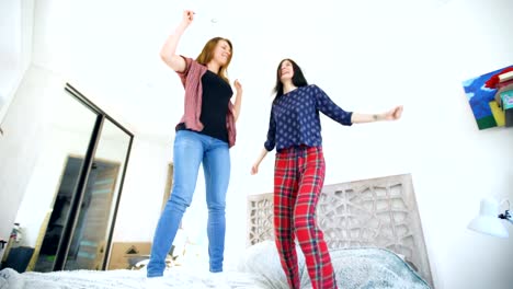 Young-happy-women-couple-dancing-over-a-bed-with-earphones-and-have-fun-in-bedroom-at-home