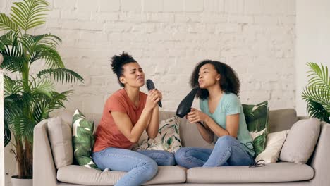 Raza-mixta-jóvenes-chicas-divertidas-bailan-cantando-con-secador-de-pelo-y-peine-sentado-en-el-sofá.-Hermanas-que-se-divierten-ocio-en-la-sala-de-estar-en-casa-concepto