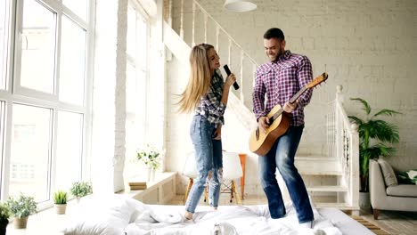 Funny-happy-and-loving-couple-dance-on-bed-singing-with-tv-controller-and-playing-guitar.-Man-and-woman-have-fun-during-their-holiday-at-home