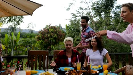 Two-Man-Taking-Food-From-Barbecue-People-Sitting-At-Table-Young-Friends-Group-Gathering-On-Summer-Terrace-Having-Party