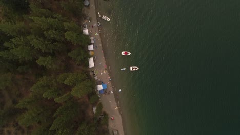 Drone-Over-Lake-Beach-Camp-Site-with-Boats-and-Tents