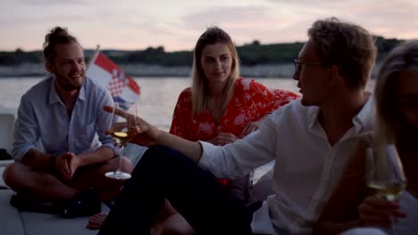 Group-of-Young-People-Talk,-Drink-Champagne-in-the-Stern-of-the-Moving-Yacht.-They-Have-Great-Vacation.-In-the-Background-Island-with-Small-Village.