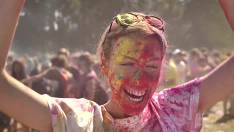 Young-happy-girl-in-colourful-powder-is-histerically-laughing-and-dancing-on-holi-festival-in-daytime-in-summer,-watching-at-camera,-color-concept,-emotional-concept