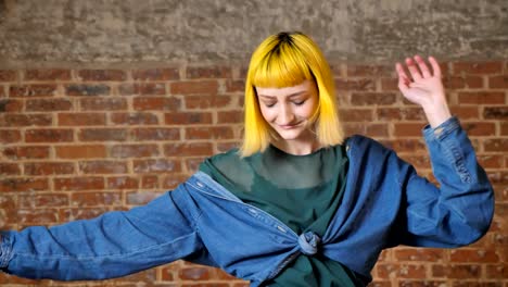 Young-unusual-woman-with-yellow-hair-dancing-and-smiling-at-camera,-isolated-over-brick-background,-happy