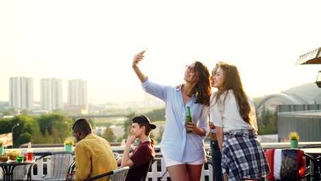 Fröhliche-Freundinnen-nehmen-Selfie-mit-Smartphone-und-lachen-stehen-auf-Dach-mit-Getränke-in-Flaschen,-die-Partei-zu-genießen.-Technologie,-Getränke--und-Jugend-Konzept.