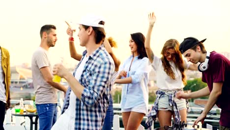 Jóvenes-están-bailando-y-riendo-mientras-hombre-DJ-está-trabajando-con-el-equipo-en-la-fiesta-en-la-azotea-en-verano.-Concepto-de-entretenimiento,-la-juventud-y-la-música.