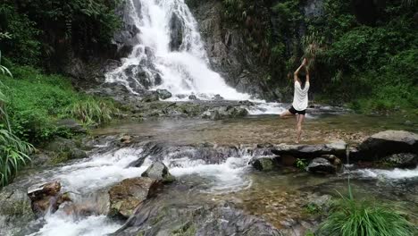 Vista-aérea-de-mujer-de-estilo-de-vida-saludable-haciendo-yoga-cerca-de-cascada-en-el-bosque