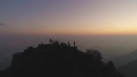 Siluetas-de-personas-en-la-parte-superior-de-la-roca-en-el-crepúsculo.-Horizonte-de-la-ciudad-en-la-niebla-con-humo.-Vista-aérea.-Drone-está-volando-hacia-arriba.-Establecimiento-de-tiro.