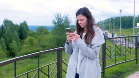 Young-brunette-woman-chatting,-typing-a-message-on-mobile-phone-walking-along-the-promenade-in-the-city.