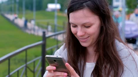 Young-brunette-woman-chatting,-typing-a-message-on-mobile-phone-walking-along-the-promenade-in-the-city.