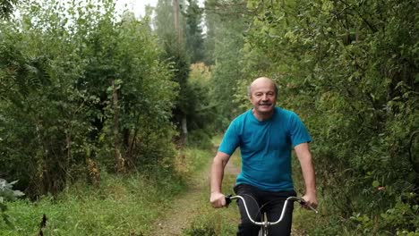 Senior-caucasian-male-in-blue-t-shirt-enjoying-his-summer-vacation-riding-a-bicycle-outdoor-between-trees.