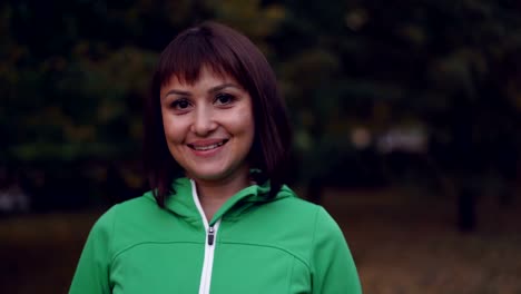 Portrait-of-pretty-young-woman-in-sportswear-looking-at-camera-and-smiling-standing-outdoors-in-park-in-autumn-evening-with-lights-visible-in-background.