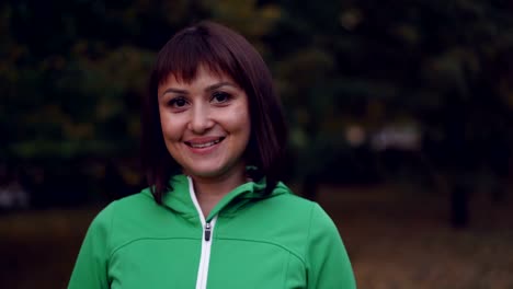 Retrato-de-una-mujer-bastante-joven-en-ropa-deportiva-mirando-a-cámara-y-sonrisa-permanente-al-aire-libre-en-el-parque-en-otoño-noche-con-luces-visibles-en-el-fondo.