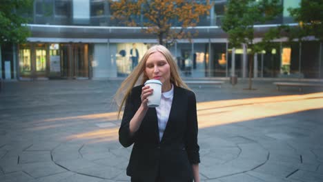 Mujer-formal-tomando-café-en-el-patio.-Elegante-mujer-rubia-en-traje-y-con-el-pelo-largo,-bebiendo-café-de-taza-blanca-en-patio-con-árboles