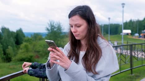 Young-brunette-woman-chatting,-typing-a-message-on-mobile-phone-walking-along-the-promenade-in-the-city.