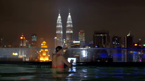 Blick-auf-Frau-im-Schwimmbad-auf-dem-Hochhaus-Dach-mit-Tablette-gegen-Nacht-Stadtlandschaft.-Kuala-Lumpur,-Malaysia