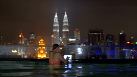 Vista-de-la-mujer-en-la-piscina-en-la-azotea-del-rascacielos-usando-tableta-contra-el-paisaje-de-la-ciudad-de-noche.-Kuala-Lumpur,-Malasia