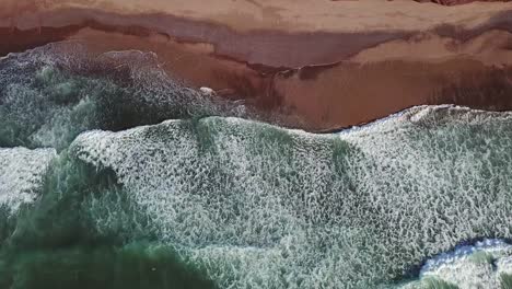 Vista-aérea-inclinación-sobre-las-olas-del-océano-y-playa-de-arena-roja