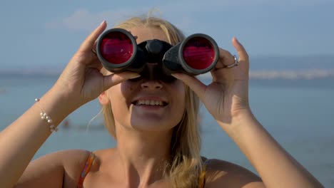 Vista-de-la-mujer-rubia-mirando-con-prismáticos-contra-mar-borrosa
