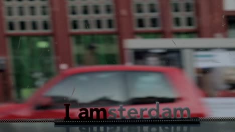 View-of-small-plastic-figure-of-Iamsterdam-letters-sculpture-on-the-bridge-against-blurred-cityscape,-Amsterdam,-Netherlands