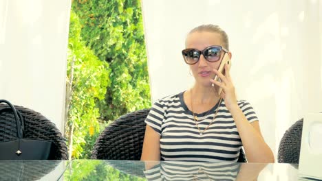 Junge-schöne-Blondine-in-Sonnenbrille-auf-dem-Smart-Phone-sitzen-auf-einem-Stuhl-im-Sommercafé-sprechen.