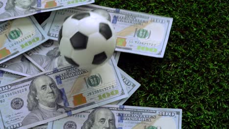 Negocio-del-balompié,-fútbol-concepto-de-juego:-un-balón-de-fútbol-en-billetes-de-un-dólar-de-los-Estados-Unidos,-que-se-extendió-sobre-la-hierba-verde.-VDO-cámara-lenta.