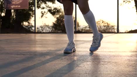Young-girl-stretching-her-legs-in-white-sneakers-and-white-golf-socks-on-the-local-basketball-court.-Unrecognizable-footage-with-sun-shining-on-the-background