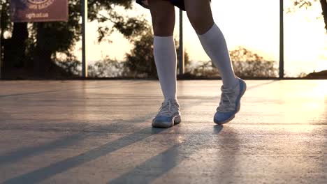 Chica-joven-que-estira-sus-piernas-en-zapatillas-blancas-y-calcetines-de-golf-blanco-en-la-cancha-de-baloncesto-local.-Material-de-archivo-irreconocible-con-el-sol-brillando-en-el-fondo