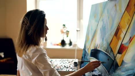 Professional-female-artist-is-working-in-studio-painting-marine-landscape-using-oil-paints-and-brush.-Beautiful-girl-is-concentrated-on-her-creative-work.