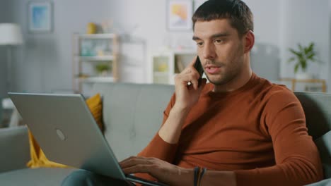 Handsome-Man-Sitting-on-Sofa-at-Home,-Talks-on-the-Mobile-Phone-and-Uses-Laptop-Computer.